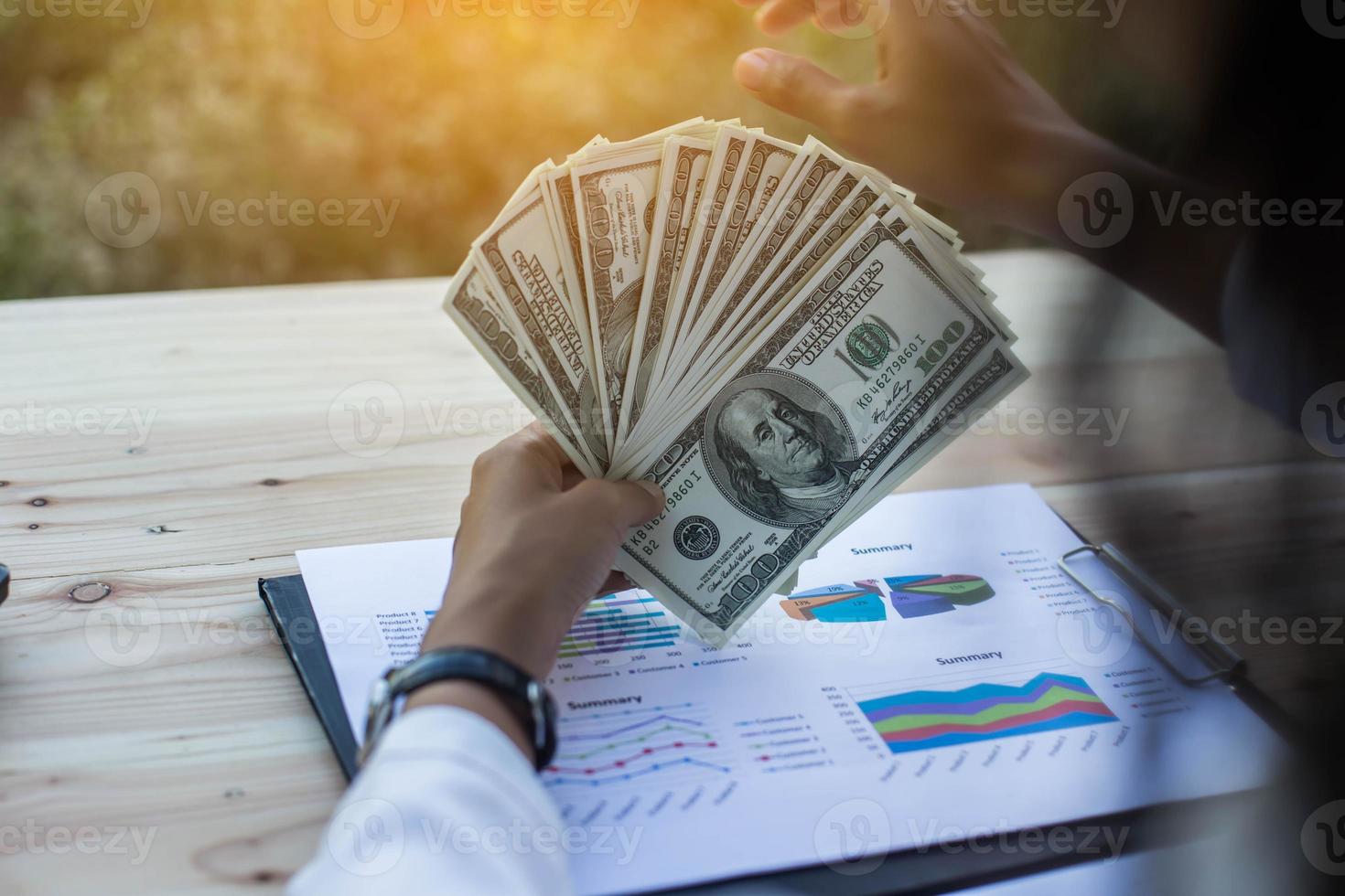 Business woman gripping The United States hundred-dollar bills in her hand and some put on the table full with business charts and notebook photo