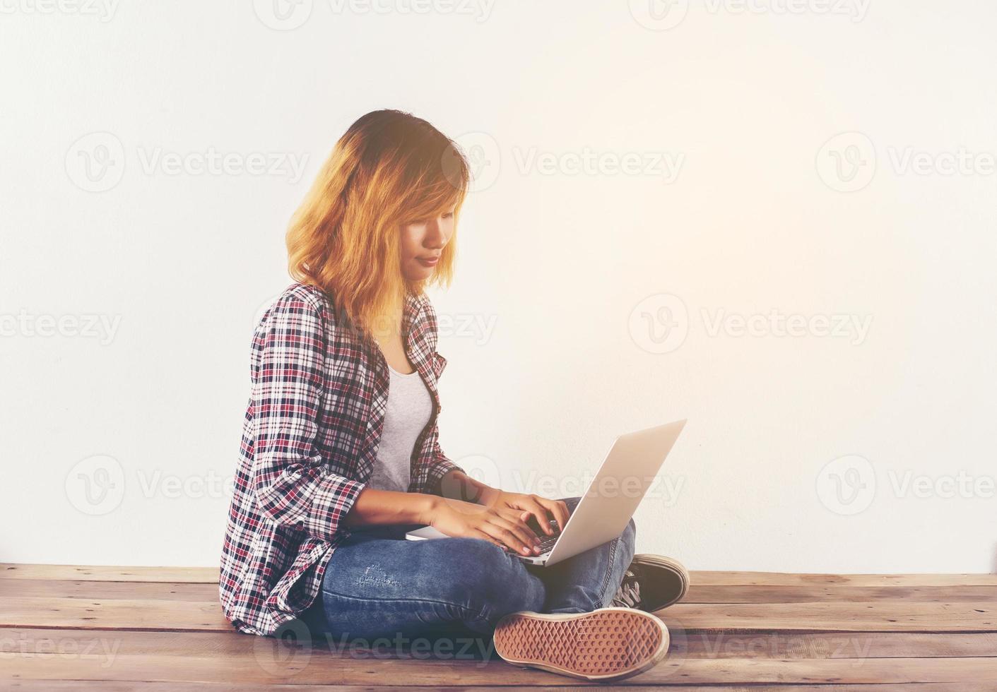 Young hipster woman sitting on wooden floor with crossed legs and using laptop on white background photo