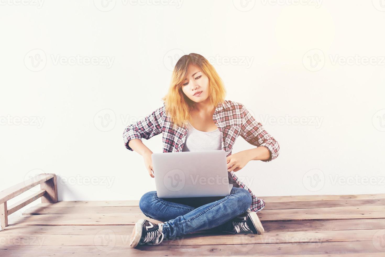 Young hipster woman sitting on wooden floor with crossed legs and using laptop on white background photo