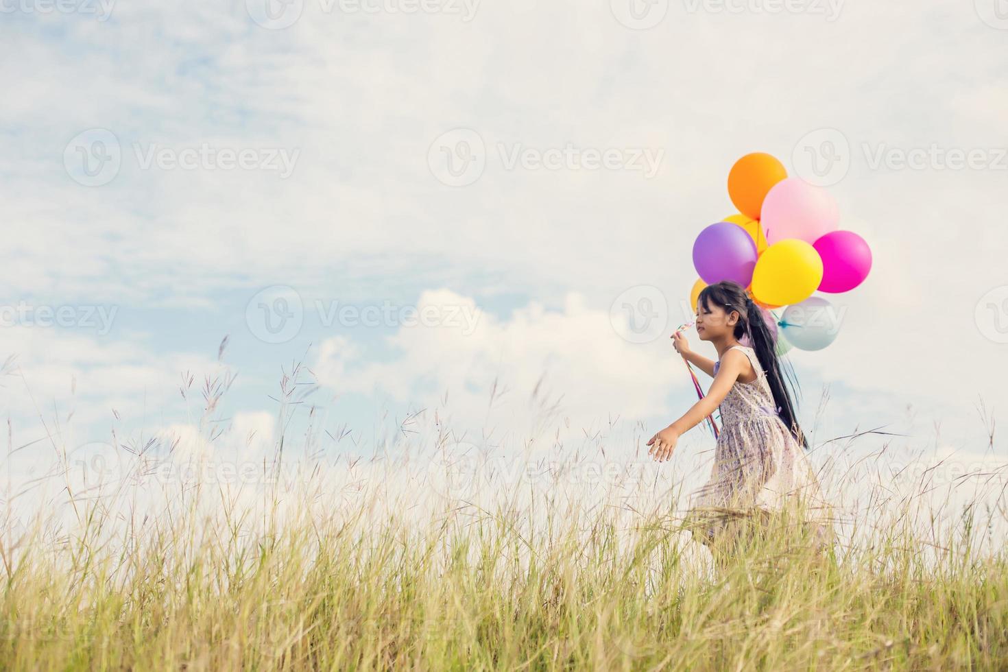 Cute little girl holding colorful balloons in the meadow against blue sky and clouds,spreading hands. photo