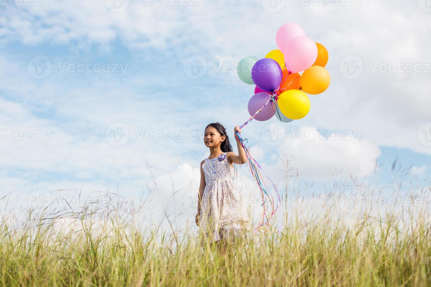 Cute little girl holding colorful balloons in the meadow against blue sky and clouds,spreading hands. photo