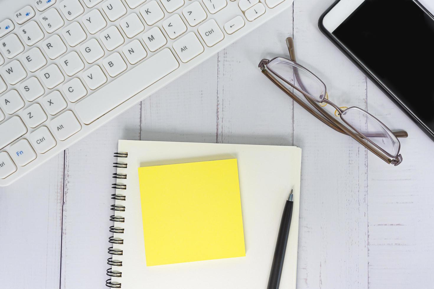 Wooden desk with notepad, computer keyboard, reading glasses, pen and smartphone photo