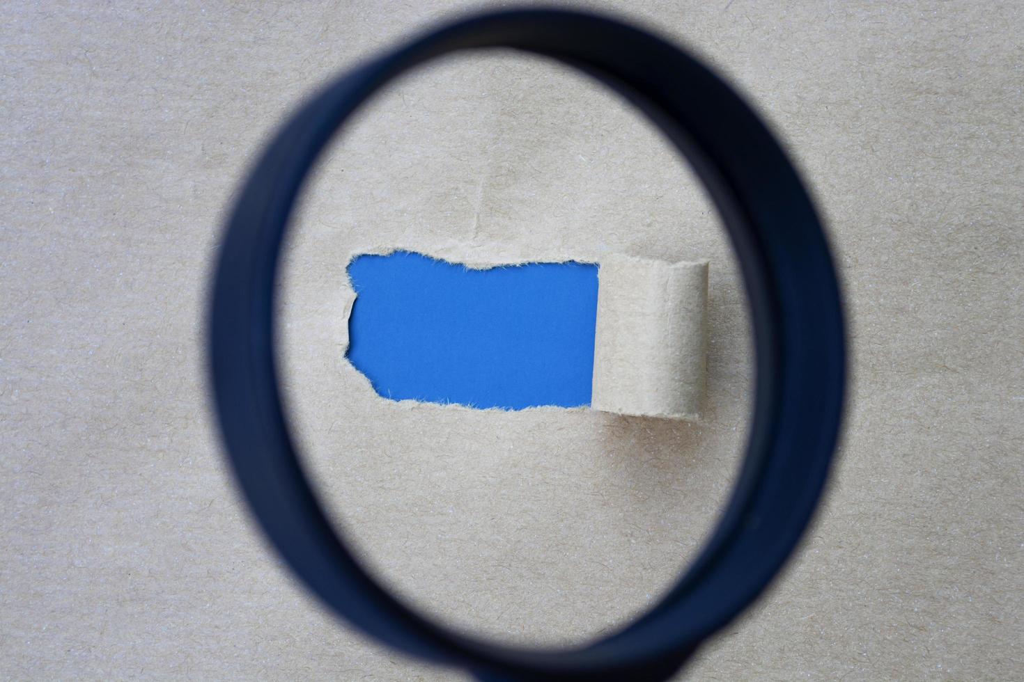 Torn hole in the sheet of brown paper on blue background with magnifying glass. photo