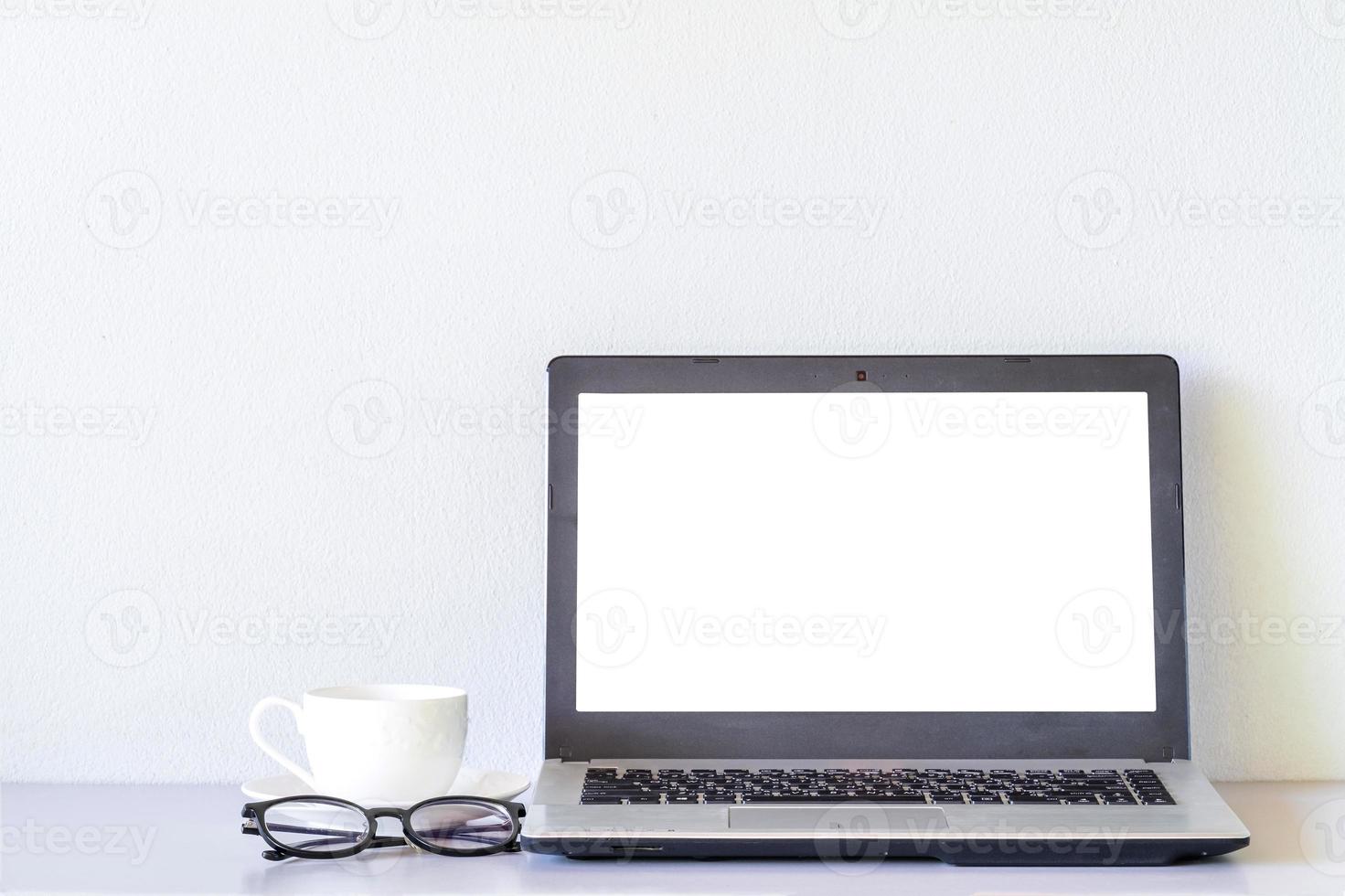 anteojos y taza de café con leche cerca de la computadora portátil se burlan de la mesa y el fondo de la pared blanca foto