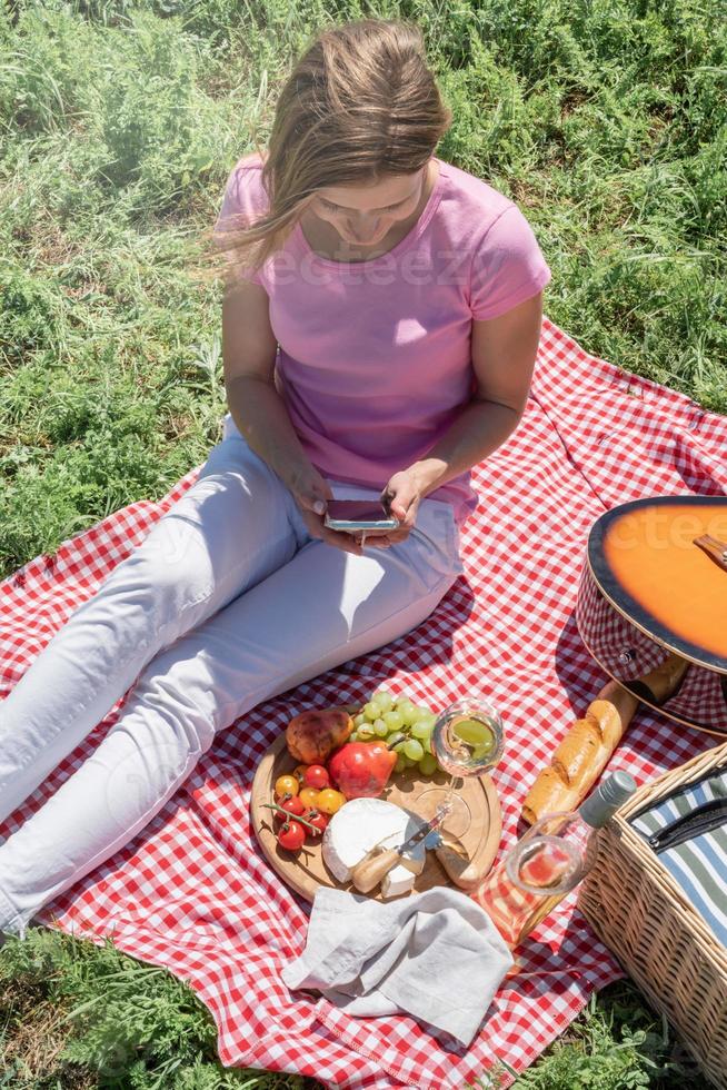 woman in white pants outside having picnic and using smartphone taking photo