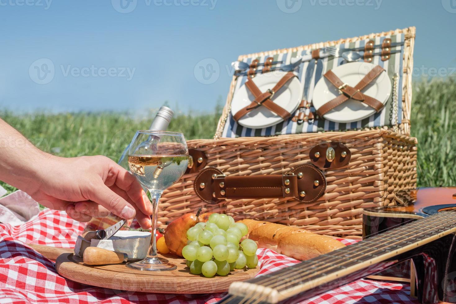 primer plano de una cesta de picnic con bebidas y comida en el césped. mano masculina sosteniendo una copa de vino foto