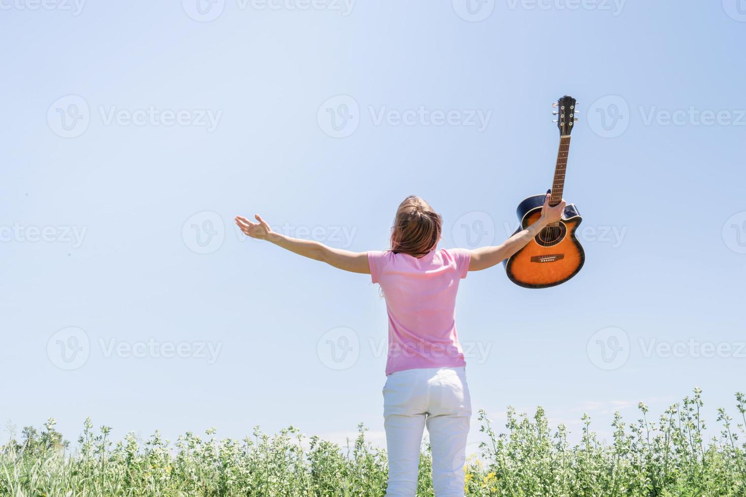 Young woman standing in grass fiel raising her hand to the sky, holding guitar photo