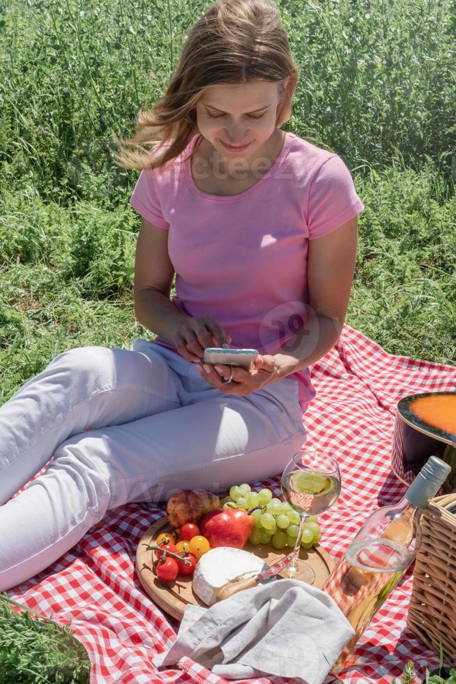 mujer con pantalones blancos afuera haciendo un picnic y usando un teléfono inteligente tomando fotos