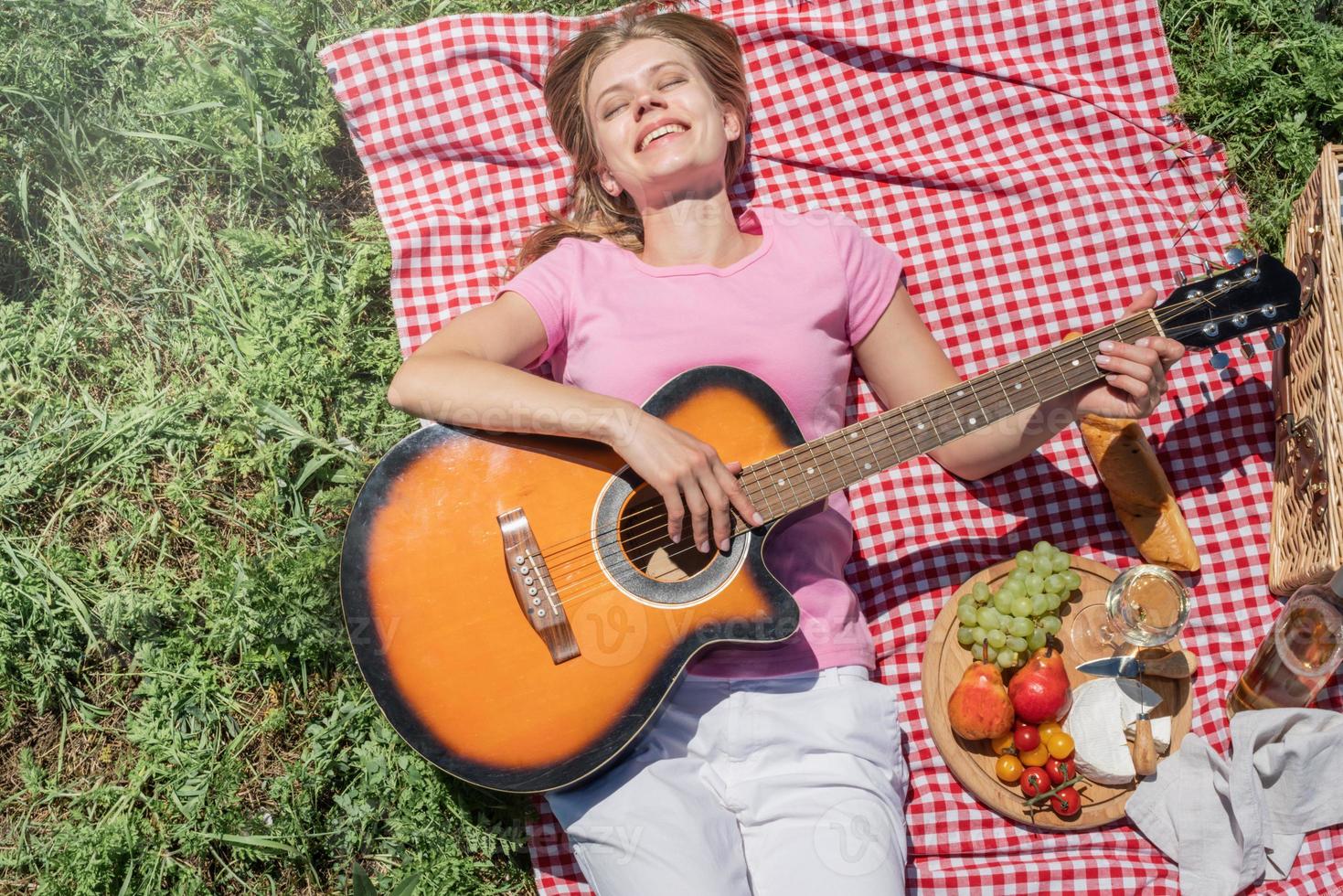 Top view of young woman in white pants outside having picnic, eating and playing guitar photo