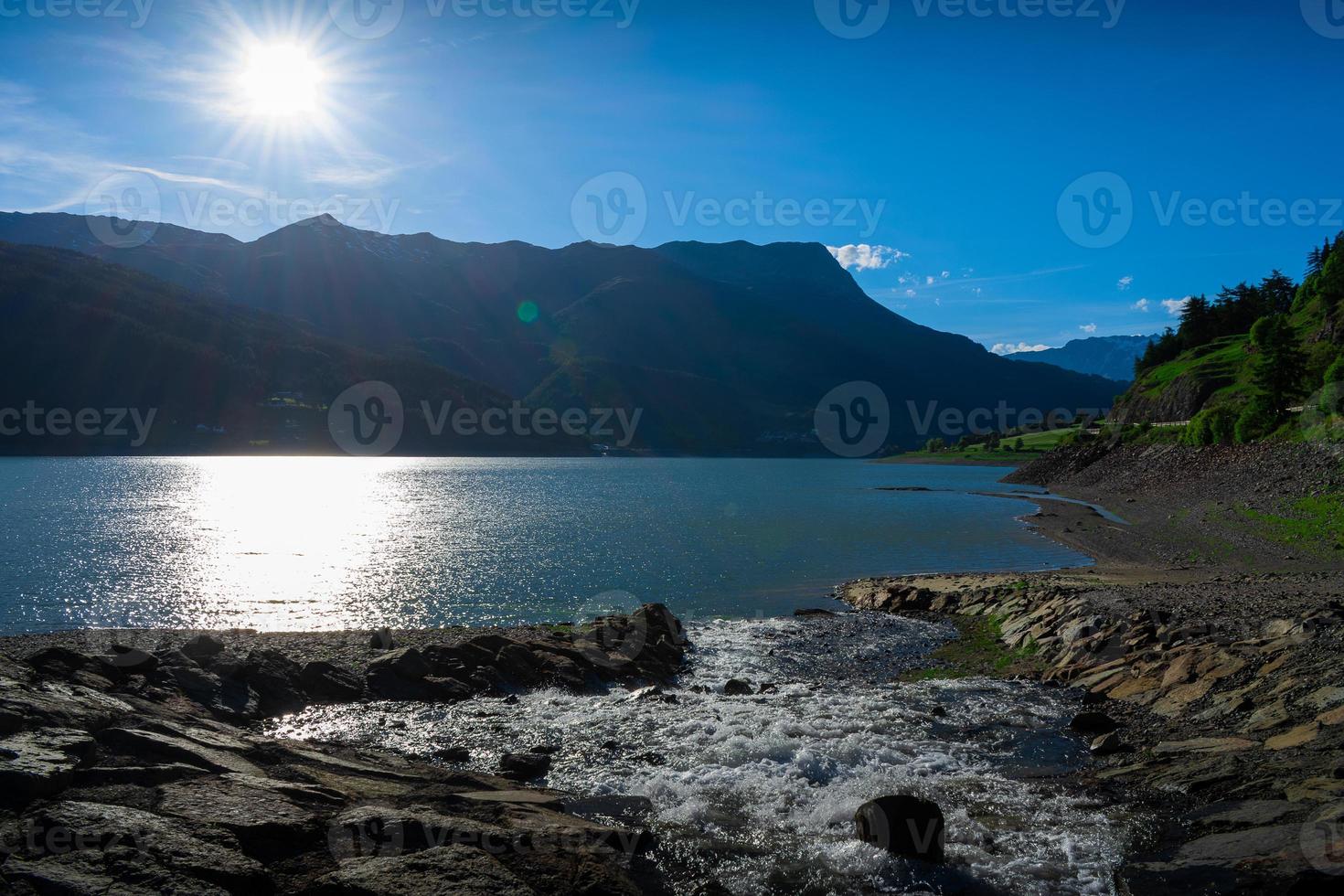 pequeña cascada en un día soleado entrando en un gran lago con montañas en segundo plano foto