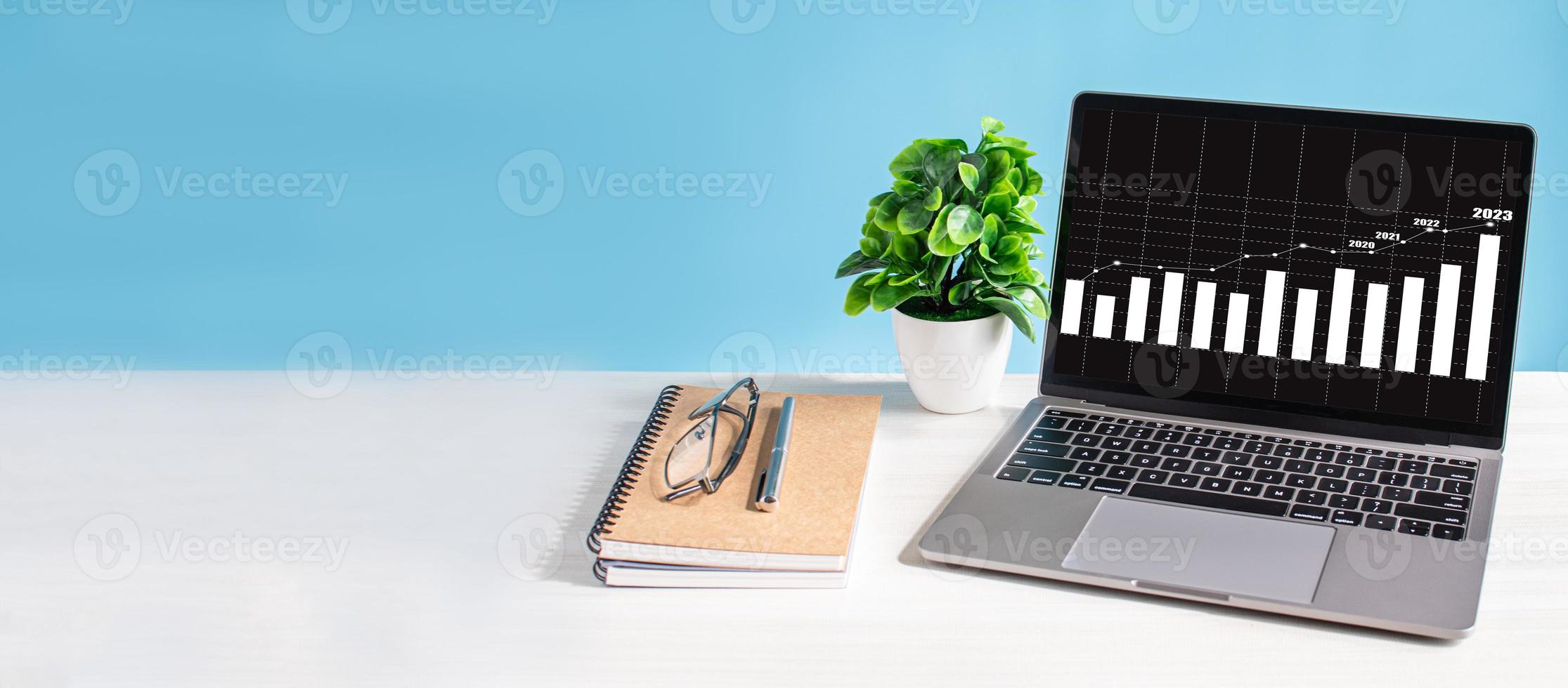 Laptop screen shows a graph of the 2023 economy rising and is placed on  wooden desk in office. Business growth concept. technology work notebook  computer network copy space, close-up, blue background 7979123