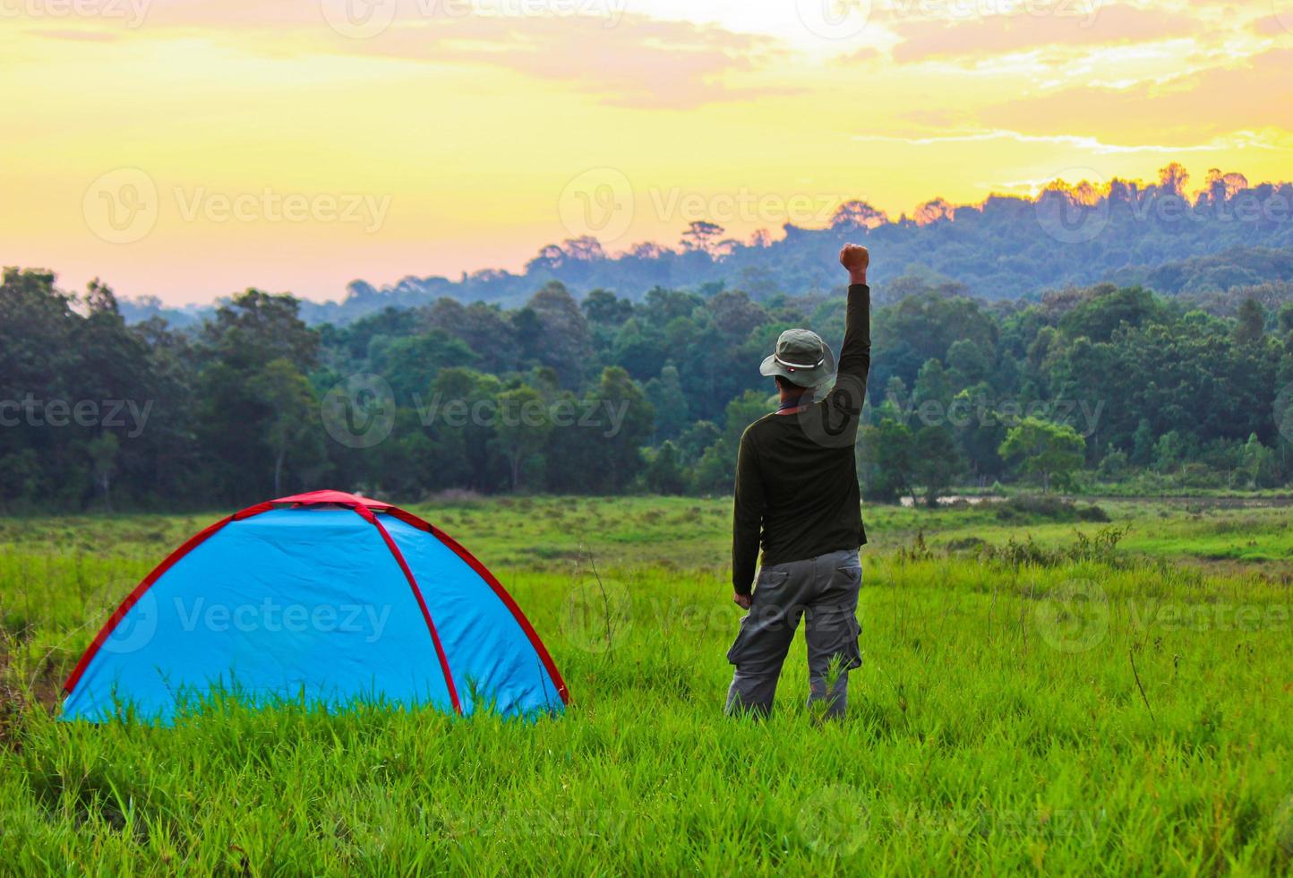 The man who stand next to the tent in the meadow put his right hand up  with trees and sky in the background photo