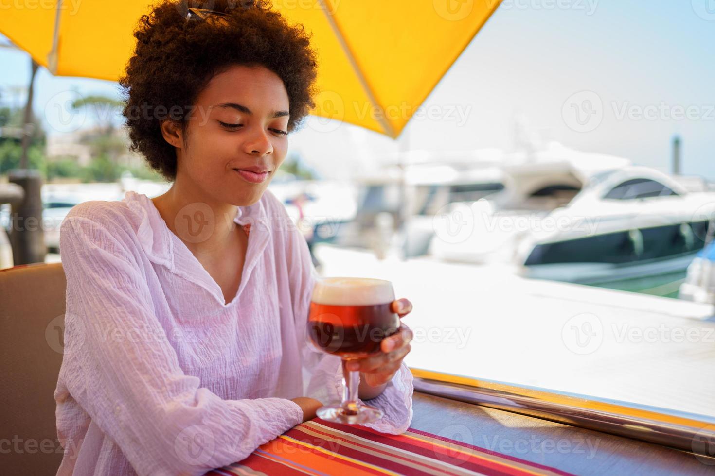 Black woman drinking a glass of cold beer with foam on the terrace of a bar in a coastal area. photo