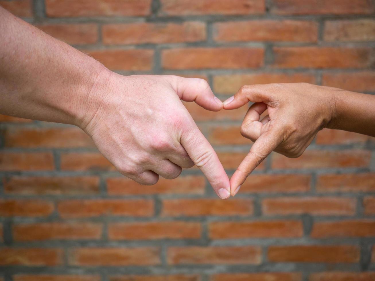 Close-up of making heart shape with hands between man and woman with brick wall background photo