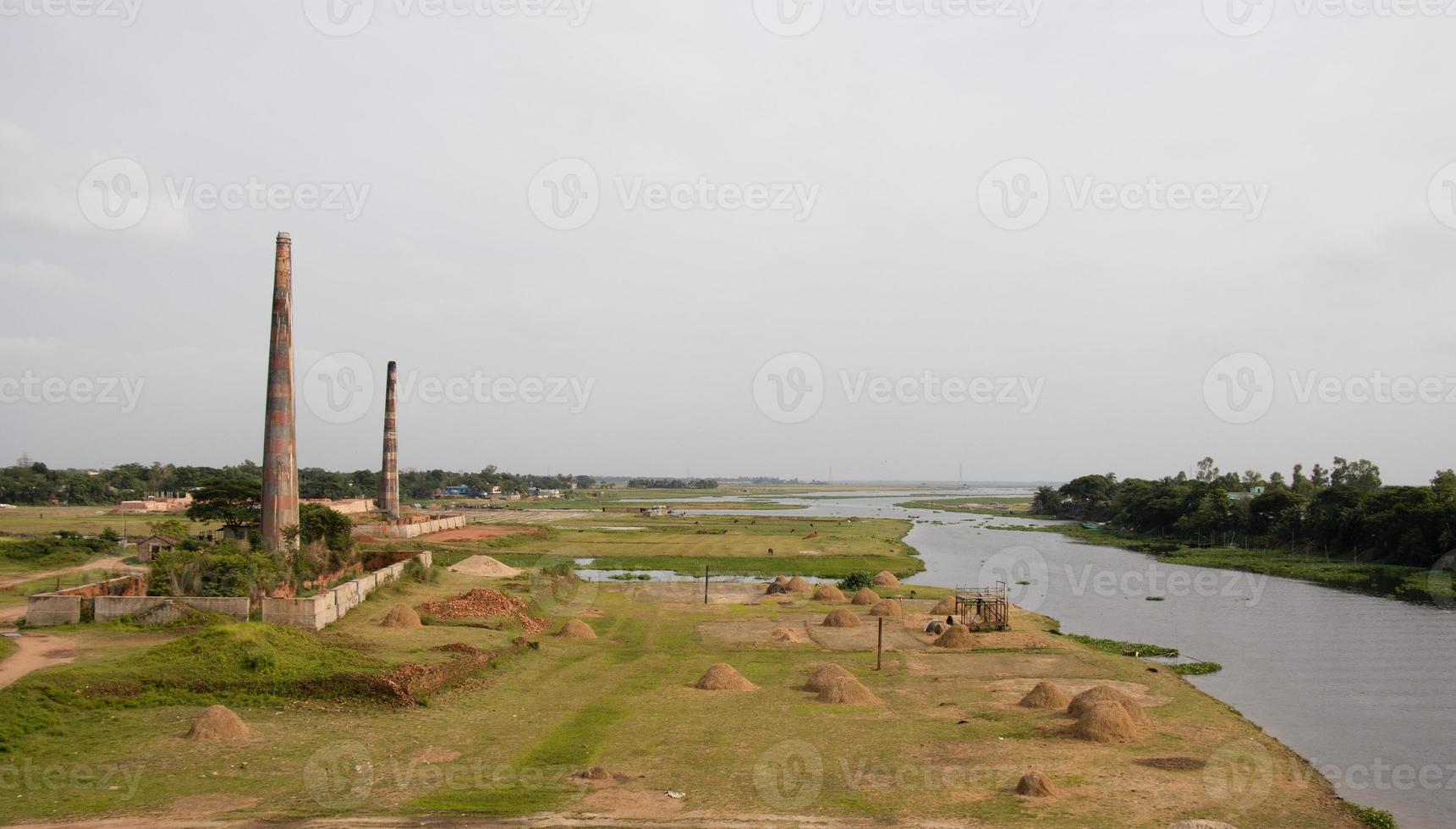 Brick kilns on a greenfield beside Titas river with a bunch of haystack photoshoots. Greenfield and tree horizon with two brick kilns. Beautiful river view with green fields and cloudy sky. photo