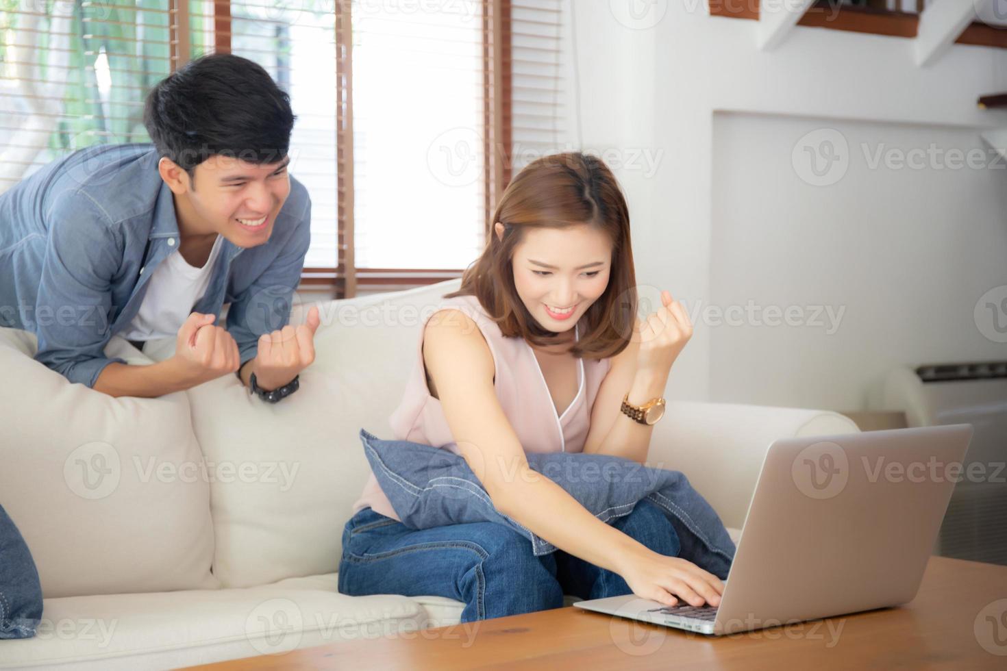 Beautiful portrait asian young couple working laptop with smile and happy sitting on couch at living room, man and woman on sofa using notebook computer with excited, business and success concept. photo