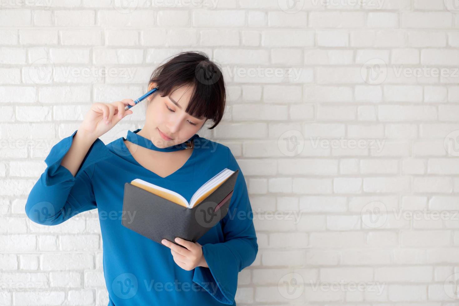 hermosa mujer asiática sonriendo de pie pensando y escribiendo un cuaderno sobre fondo blanco de cemento de hormigón en casa, tarea de niña en concepto de libro, educación y estilo de vida. foto