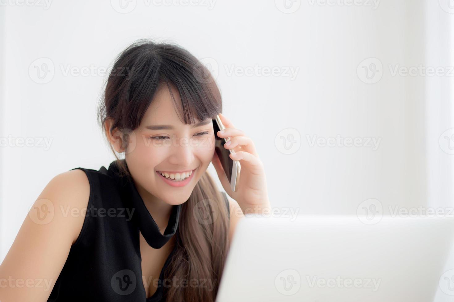 Beautiful young freelance asian woman smiling working on laptop computer and talking mobile smart phone at desk office with professional, girl using notebook, business and lifestyle concept. photo