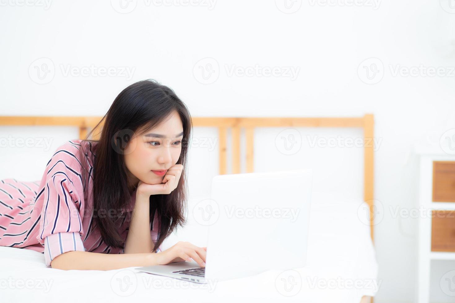 Beautiful asian young woman lying on bed using laptop at bedroom for leisure and relax, freelance with girl working notebook, communication concept. photo