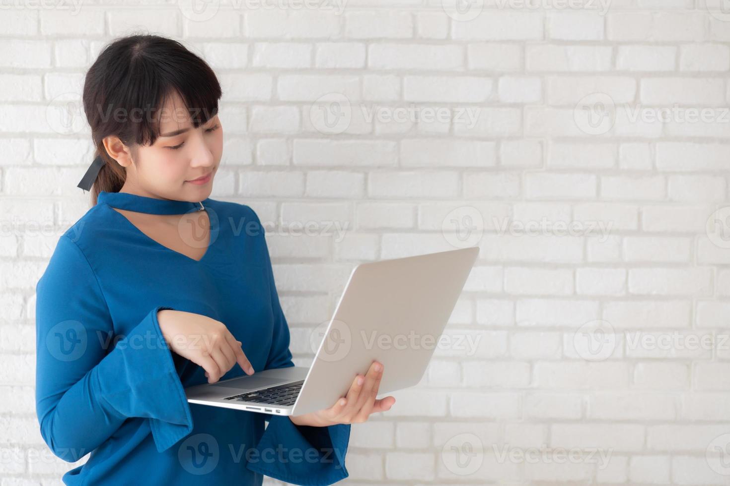 Beautiful portrait asian young woman smile using laptop standing at workplace on cement concrete background, girl happy with computer internet online, lifestyle and freelance business concept. photo