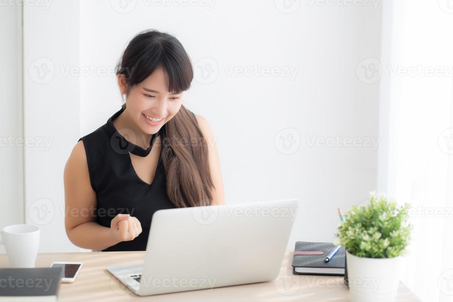 Beautiful portrait asian young woman smile working online laptop computer with happy satisfied sitting on desk office, girl using notebook with excited and glad, business and success concept. photo