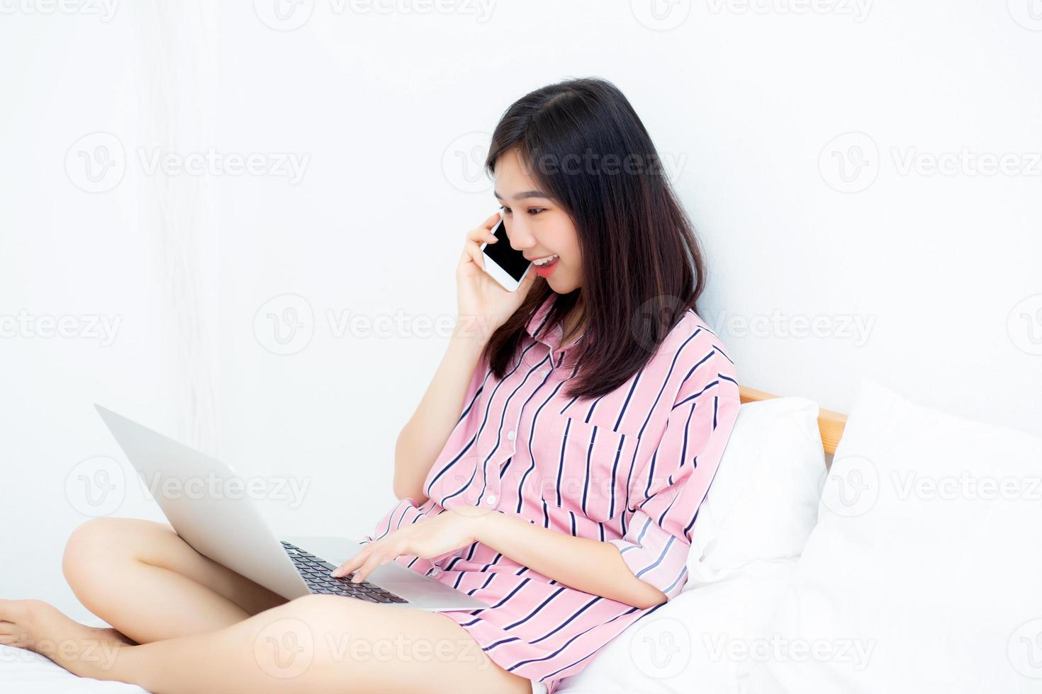 Asian woman setting on bed using laptop computer and talk smart phone at bedroom for leisure and relax, freelance with girl working notebook and calling telephone, communication and lifestyle concept. photo