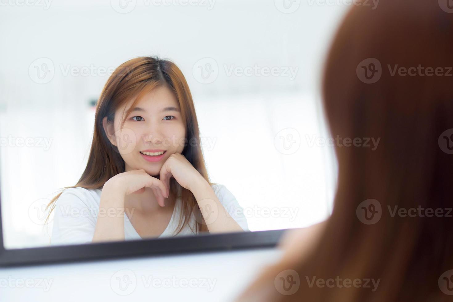 Beautiful of portrait asian young woman examining with face and smile looking on mirror at bedroom, girl beauty of makeup and dressed up with reflection, lifestyle and wellness concept. photo