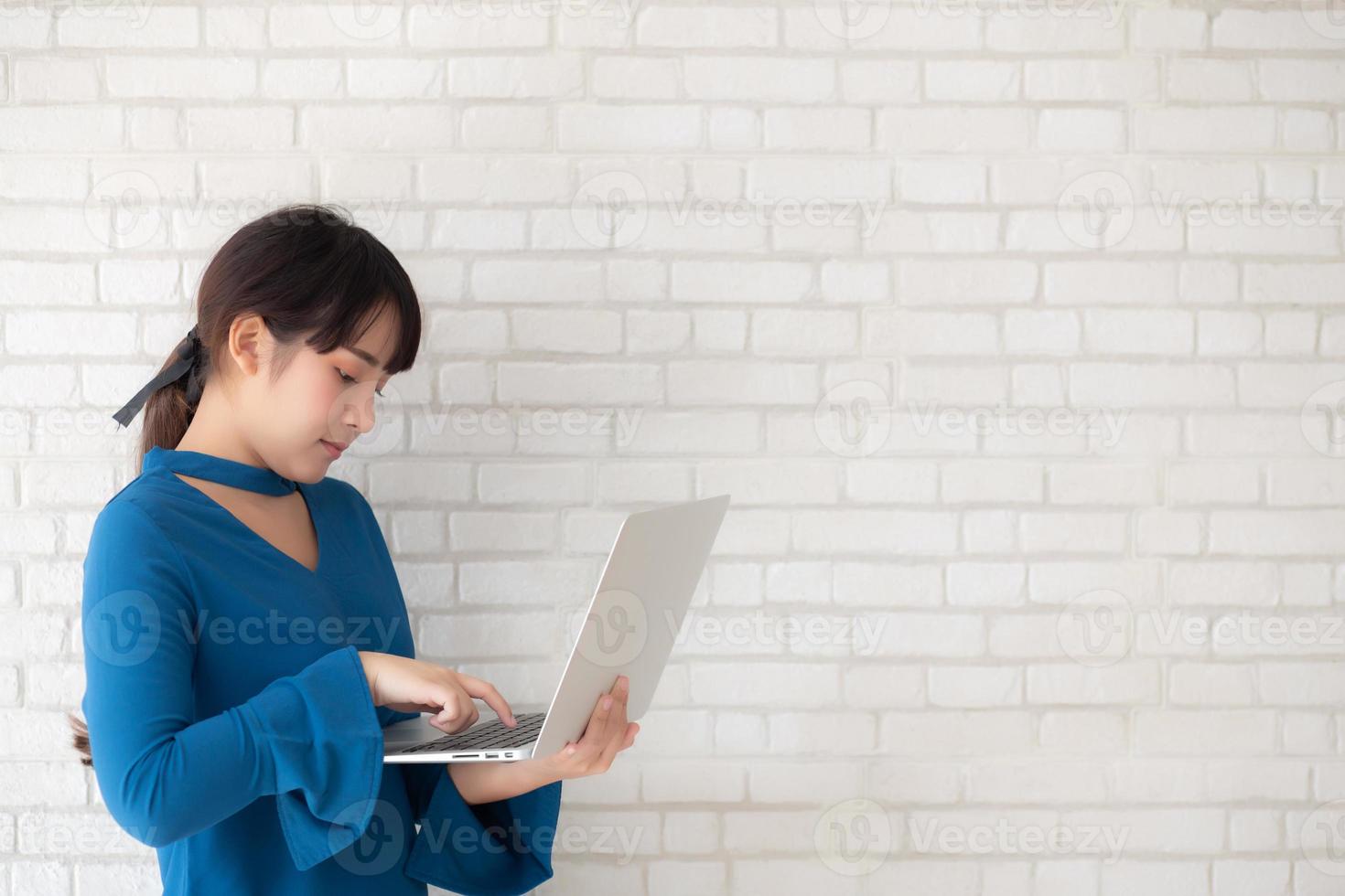 Beautiful portrait asian young woman smile using laptop standing at workplace on cement concrete background, girl happy with computer internet online, lifestyle and freelance business concept. photo