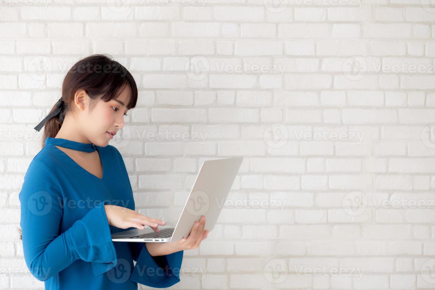 Beautiful portrait asian young woman smile using laptop standing at workplace on cement concrete background, girl happy with computer internet online, lifestyle and freelance business concept. photo