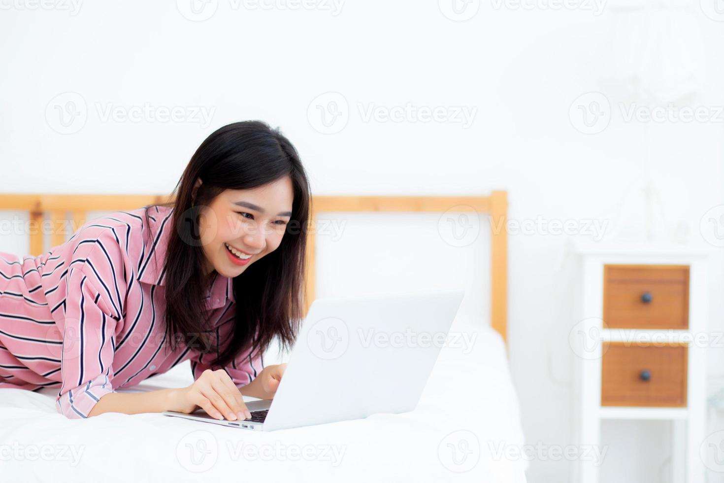 Beautiful asian young woman lying on bed using laptop at bedroom for leisure and relax, freelance with girl working notebook, communication concept. photo