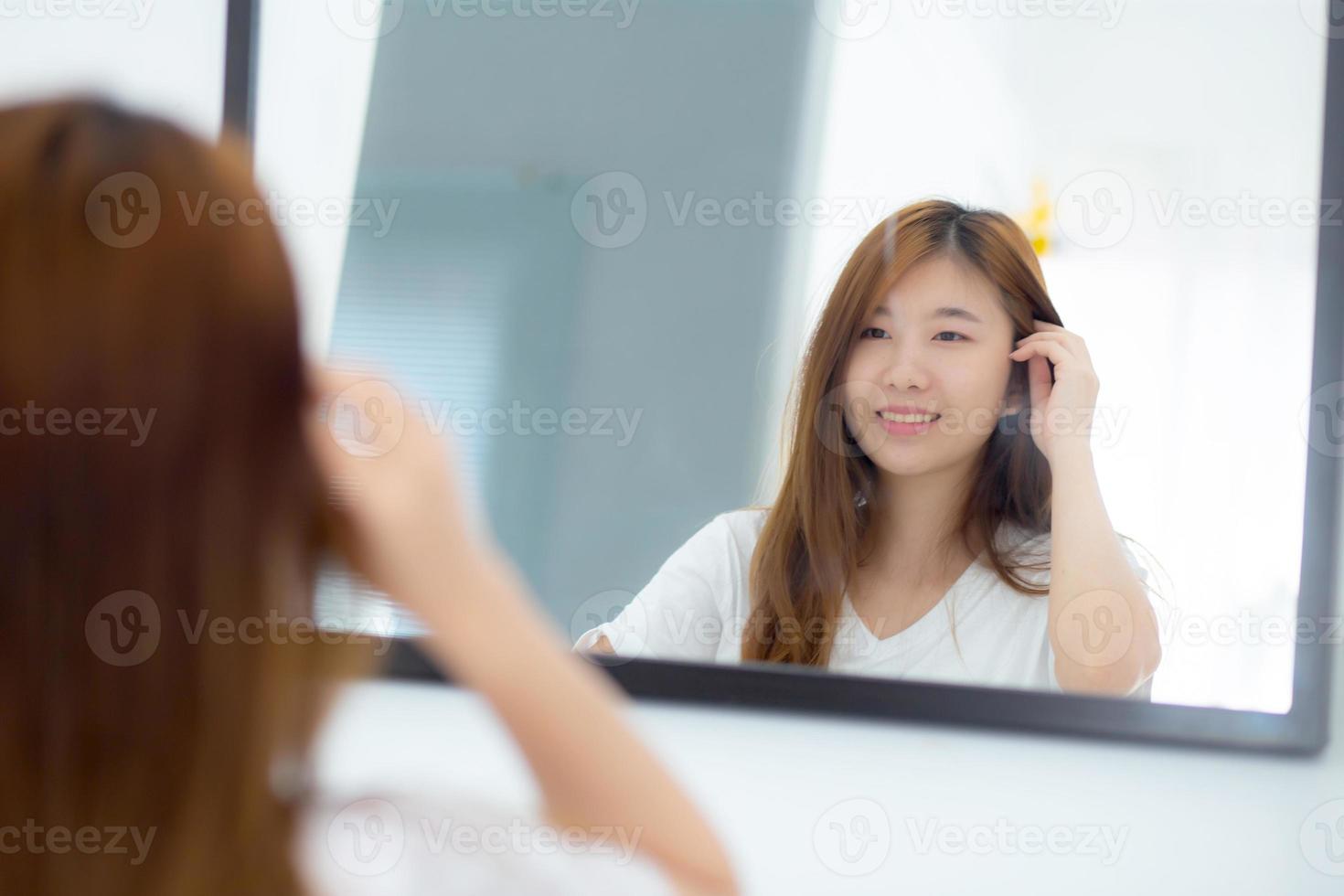 Beautiful of portrait asian young woman examining with face and smile looking on mirror at bedroom, girl beauty of makeup and dressed up with reflection, lifestyle and wellness concept. photo