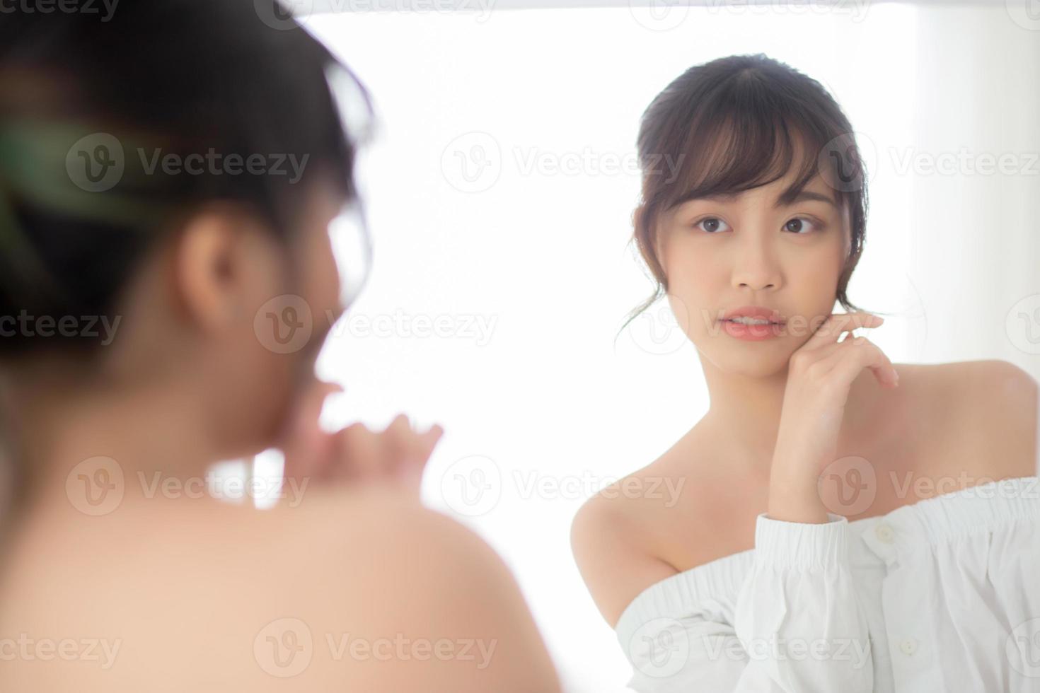 Beauty portrait young asian woman smiling look at mirror of checking skin care caucasian with wellness in the bedroom, beautiful girl happy touching face in reflection for health, lifestyle concept. photo