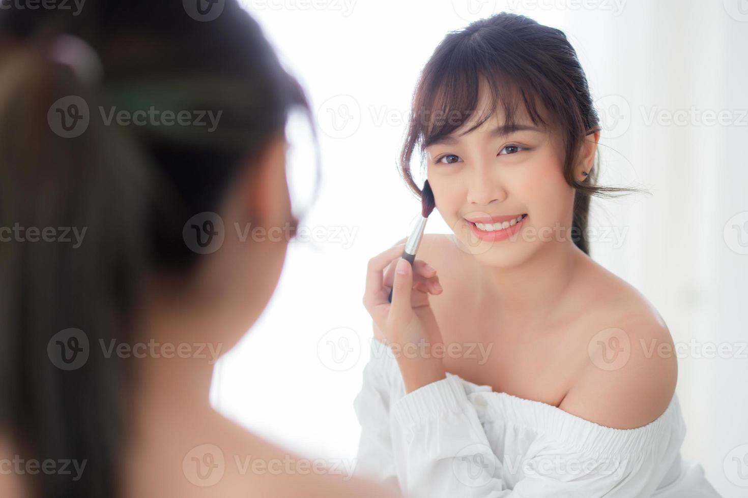 retrato de belleza mujer asiática joven sonrisa con cara mirando espejo aplicando maquillaje con cepillo de mejilla en el dormitorio, hermosa chica sosteniendo colorete, cuidado de la piel y concepto cosmético. foto