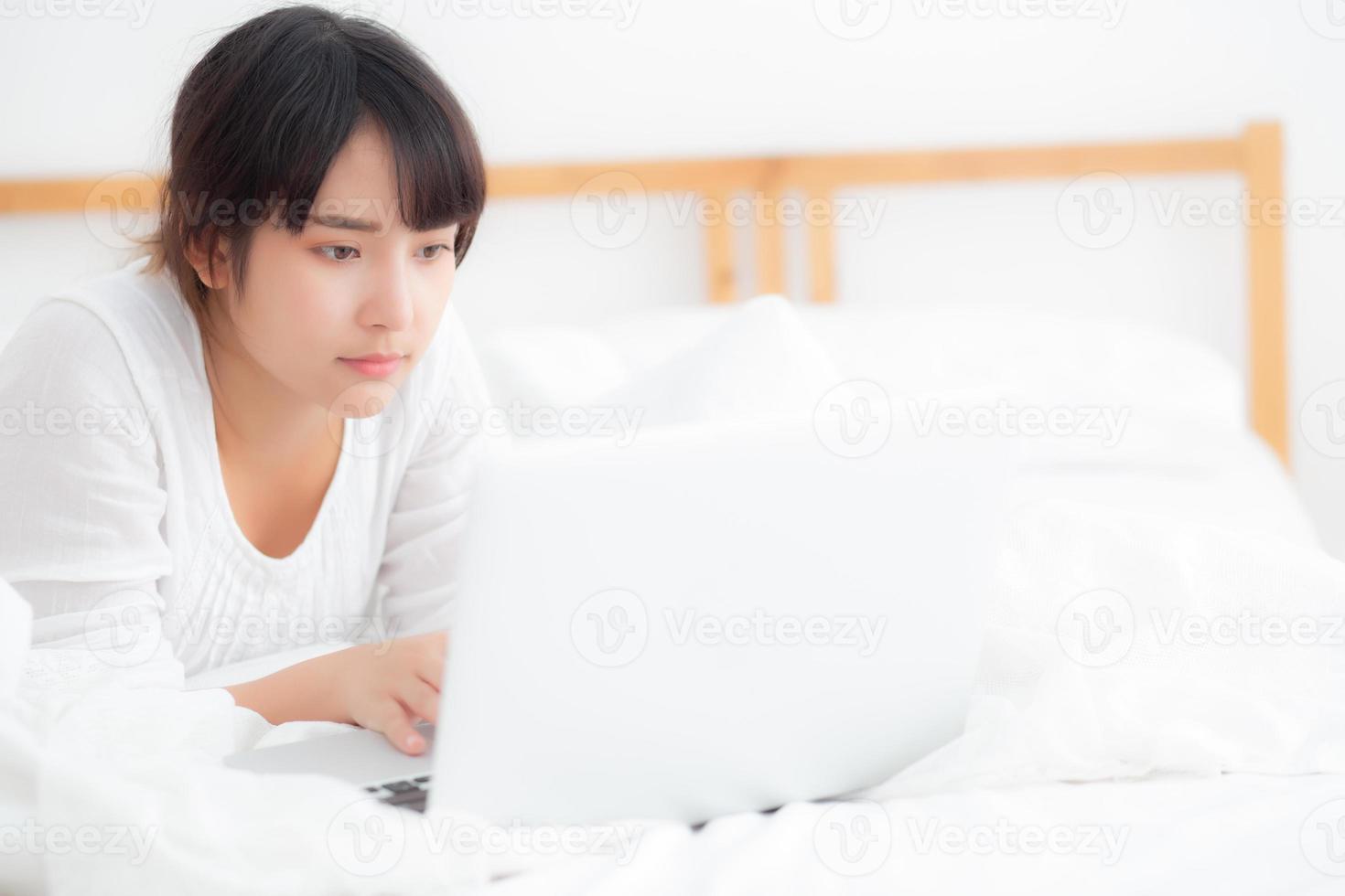Beautiful portrait asian young woman smile lying on bed using laptop computer with happy at bedroom for leisure and relax, freelance with girl working notebook, communication concept. photo