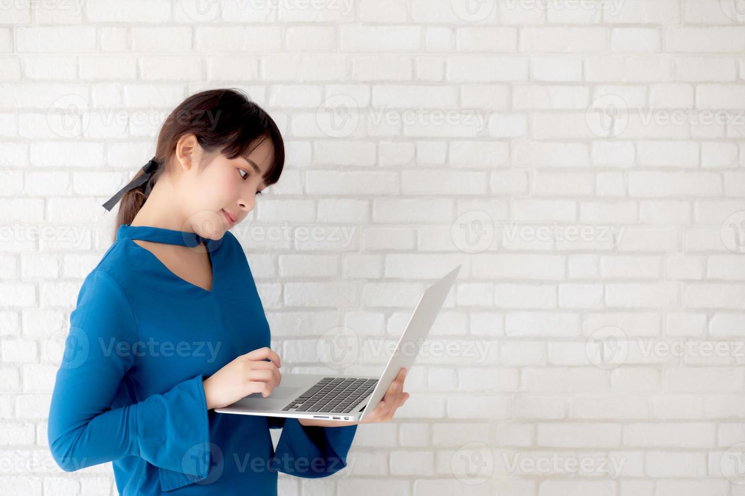 Beautiful portrait asian young woman smile using laptop standing at workplace on cement concrete background, girl happy with computer internet online, lifestyle and freelance business concept. photo