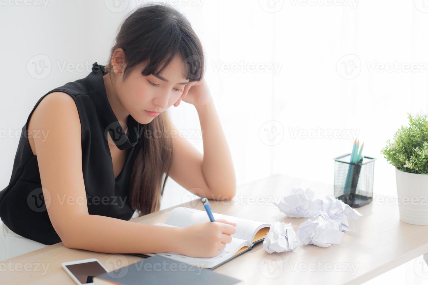 hermosa mujer asiática cansada y estresada por escribir con exceso de trabajo en el escritorio, chica preocupada sin idea con cuaderno y papel arrugado en la oficina, concepto independiente y de negocios. foto