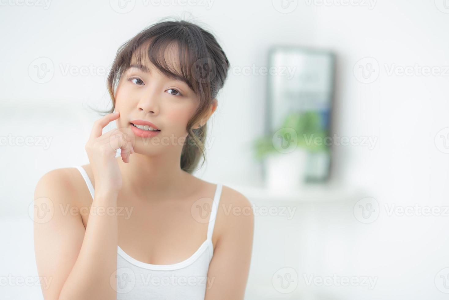 retrato hermosa mujer asiática maquillaje de cosmética, chica mano toque mejilla y sonrisa atractiva, cara belleza perfecta con bienestar en el dormitorio en casa con cuidado de la piel y concepto de salud. foto