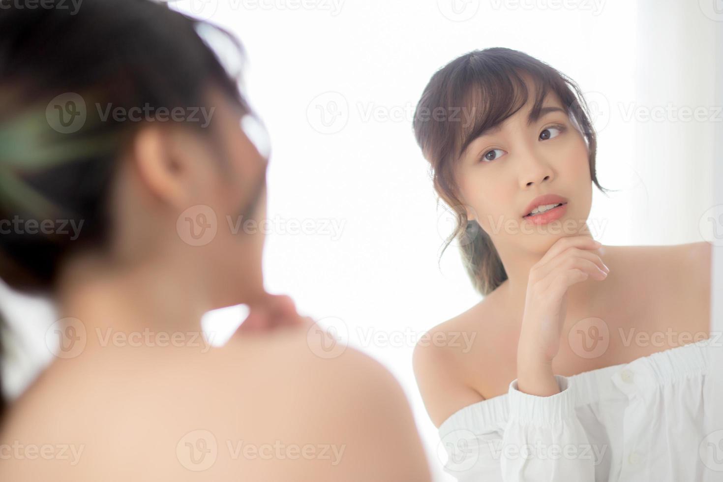 Beauty portrait young asian woman smiling look at mirror of checking skin care caucasian with wellness in the bedroom, beautiful girl happy touching face in reflection for health, lifestyle concept. photo