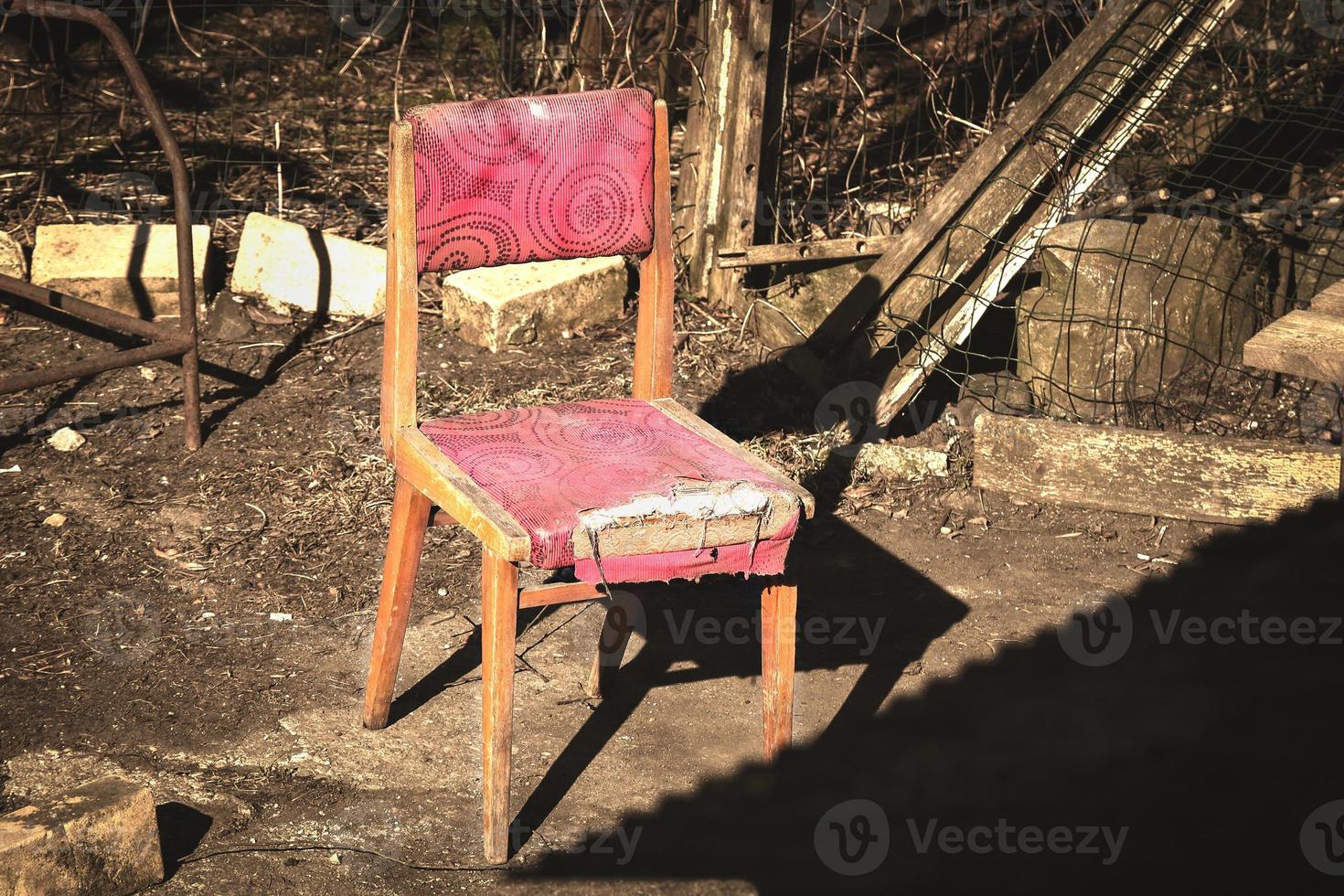 Old wooden chair with a torn red seat fabric standing outdoors in bright evening sunlight with long shadow photo