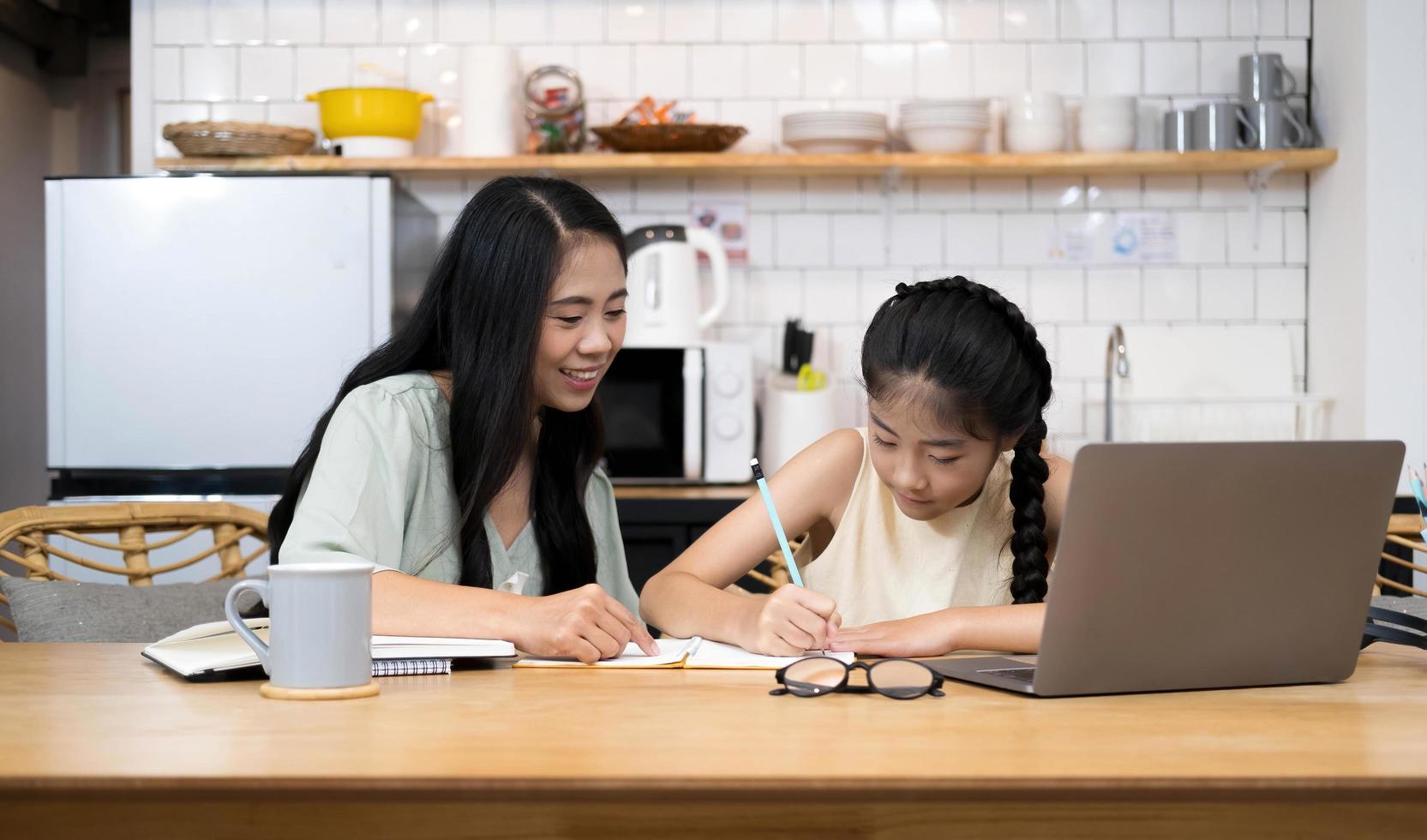 Mother and asian kid little girl learning on laptop computer making homework studying knowledge with online education e-learning system. children video conference with teacher tutor at home photo