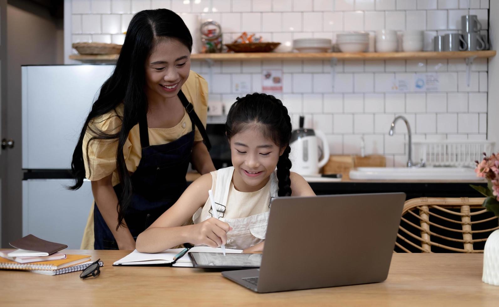 Mother and asian kid little girl learning and looking at laptop computer making homework studying with online education e-learning system.children video conference with teacher tutor at home photo