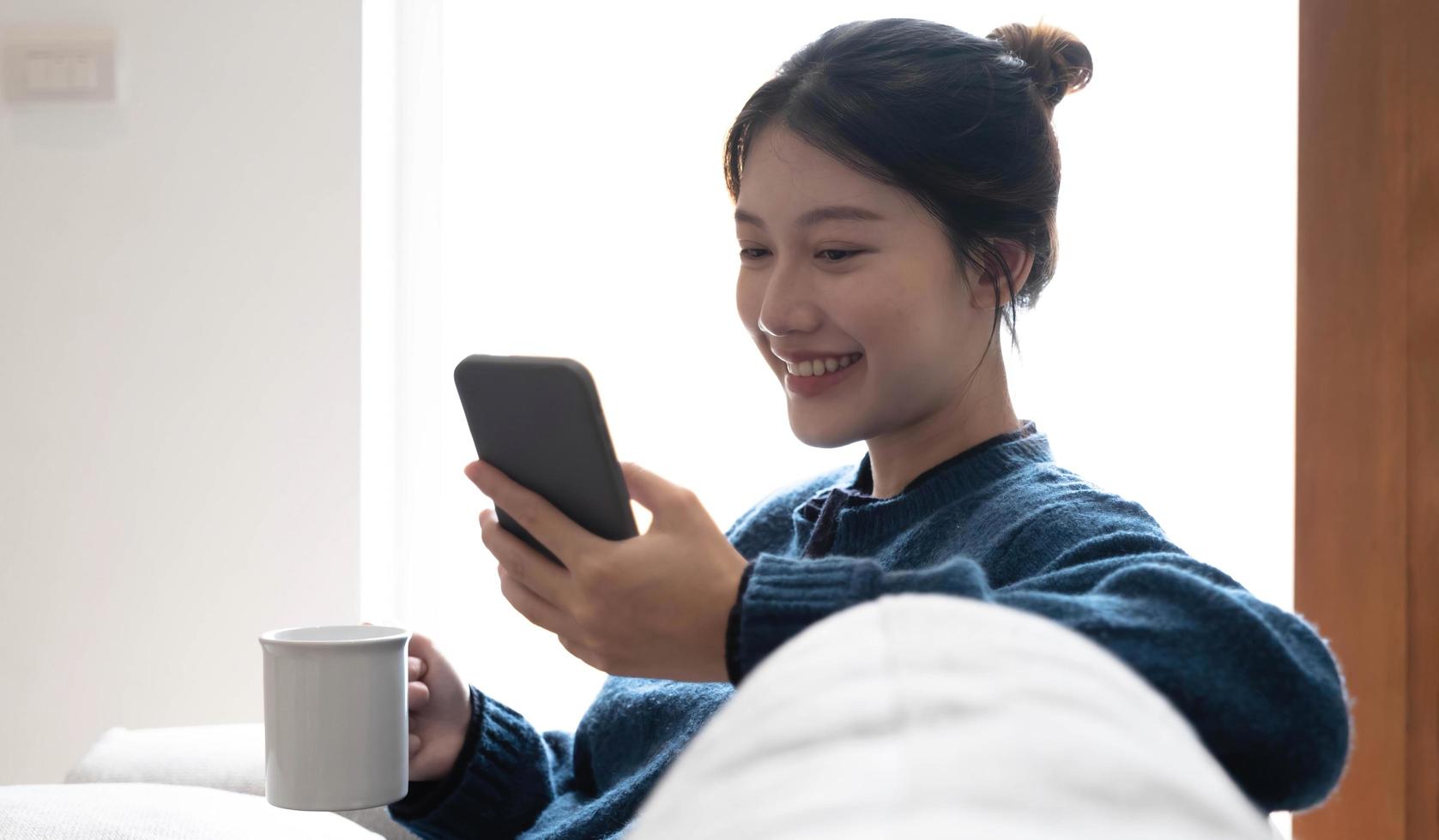 Portrait of a smiling asian women sitting at sofa and drink coffee looking at smartphone. photo