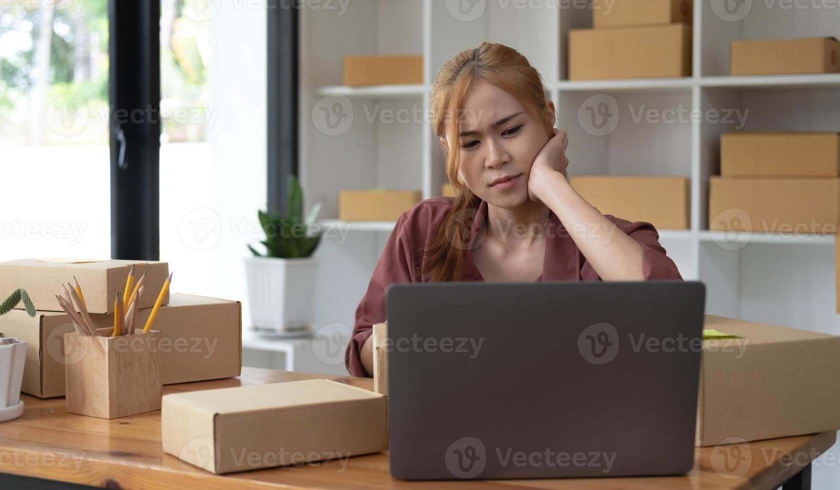 A portrait of young serious Asian woman working with laptop in the office full of packages and boxes stacking up, busy looking table, for SME, delivery, start up business and home office concept. photo