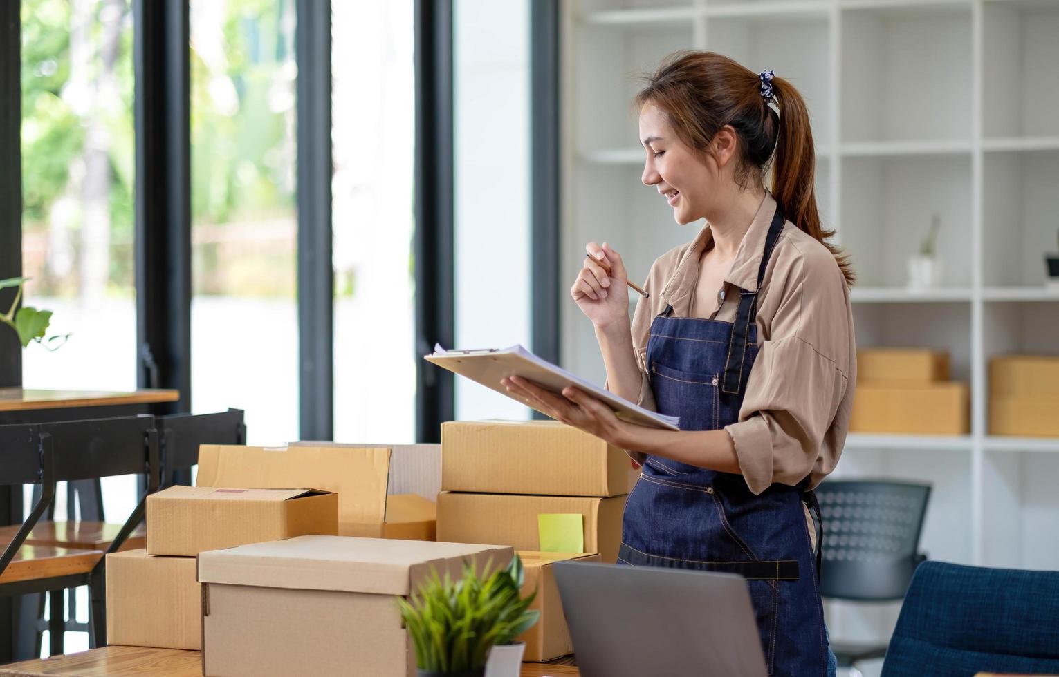 Beautiful smart Asian young entrepreneur business woman owner of SME checking product on stock and write on clipboard working at home. Small business owner at home office concept. photo