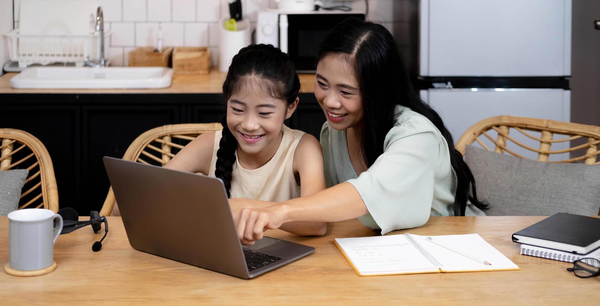 Mother and asian kid little girl learning on laptop computer making homework studying knowledge with online education e-learning system. children video conference with teacher tutor at home photo