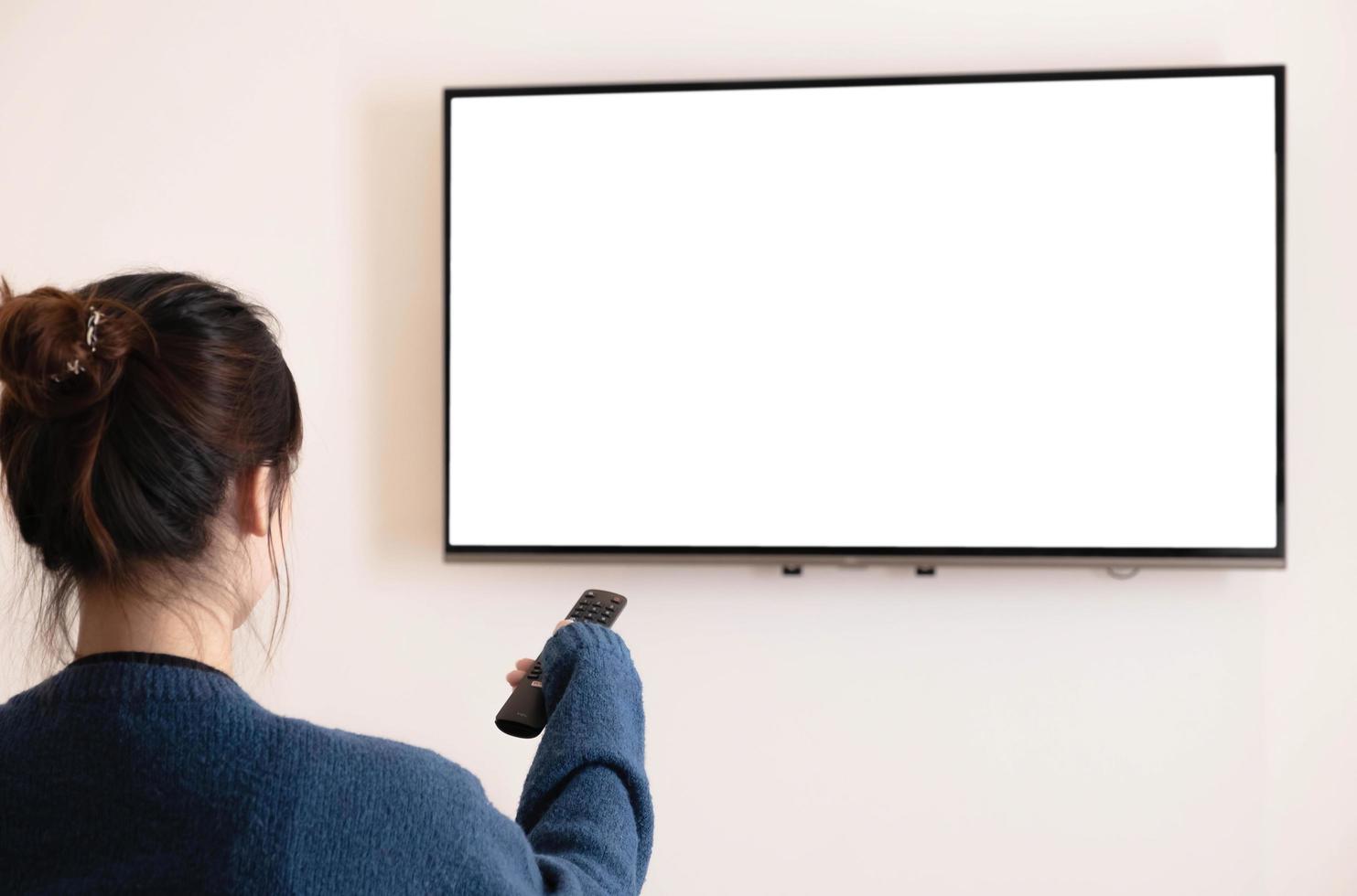 detrás de una joven asiática viendo televisión de pantalla blanca en el sofá y usando el control remoto en casa foto