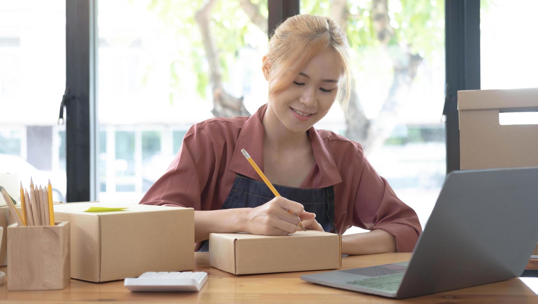 Close up woman sitting in the office full of packages in the background emplaning on a laptop and using a calculator, for online business, SME, finance, e-commerce and delivery concept. photo