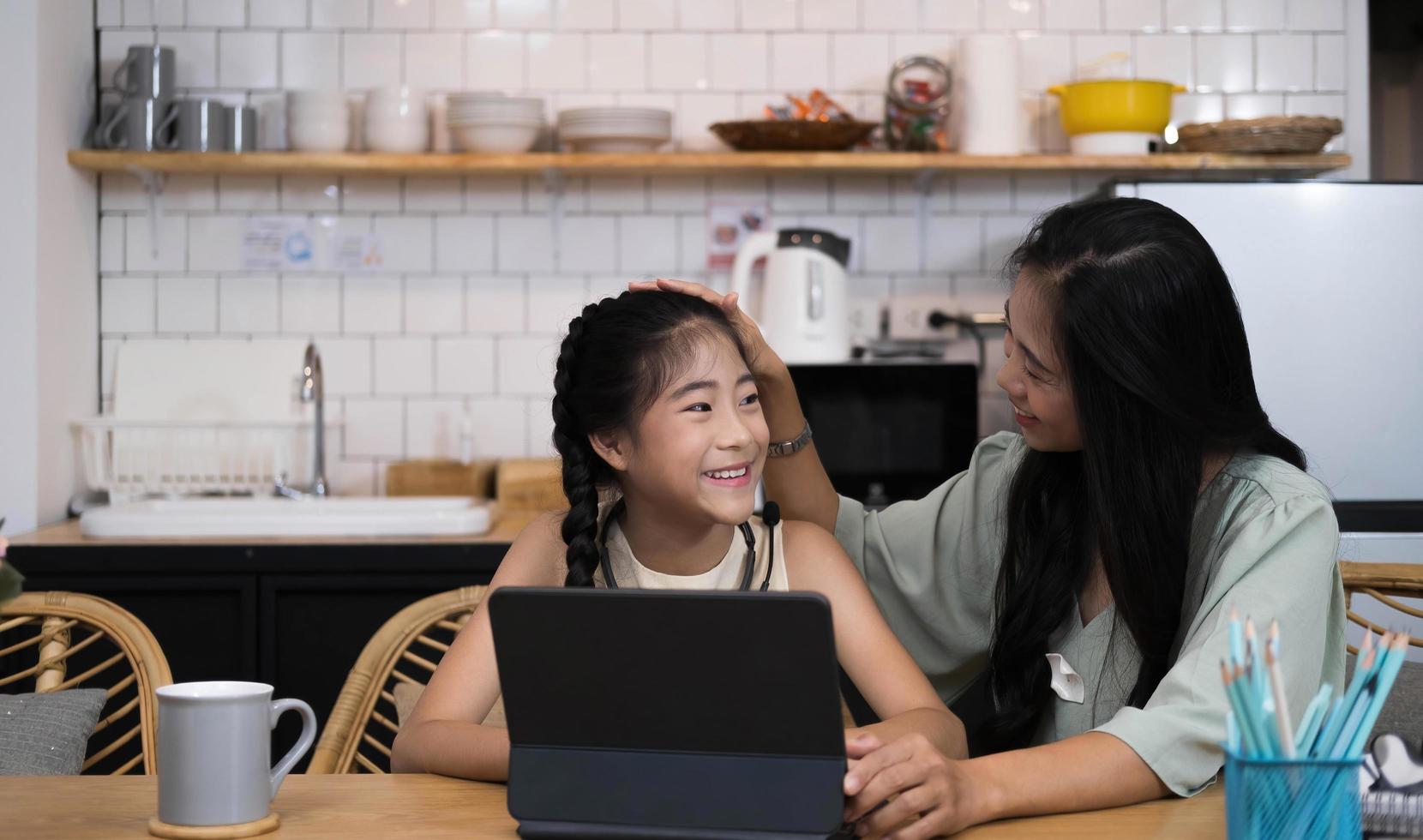 Mother and asian kid little girl learning and looking at laptop computer making homework studying knowledge with online education e-learning system.children video conference with teacher tutor at home photo