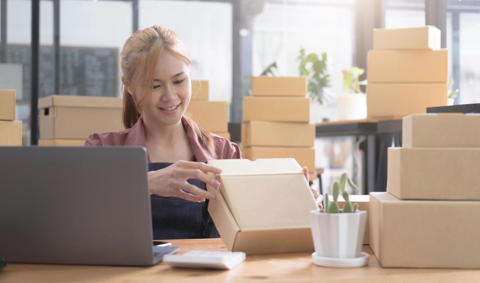 Happy young asian woman startup small business freelance holding parcel box and computer laptop and sitting on chair, Online marketing packing box delivery concept photo