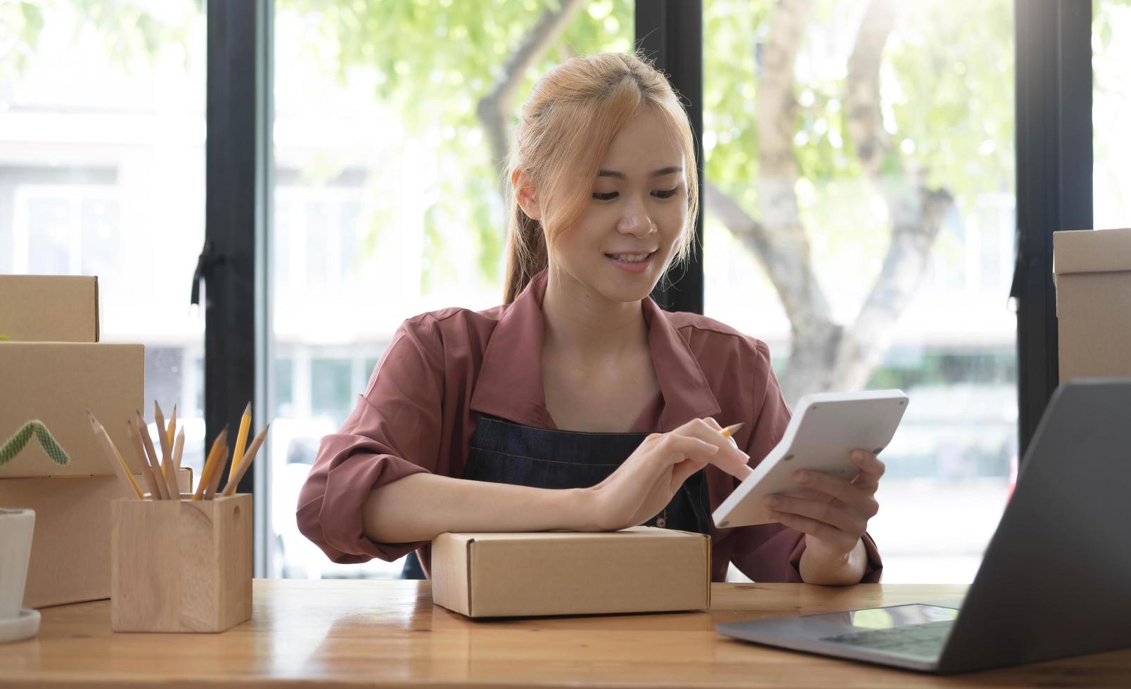 Close up woman sitting in the office full of packages in the background emplaning on a laptop and using a calculator, for online business, SME, finance, e-commerce and delivery concept. photo